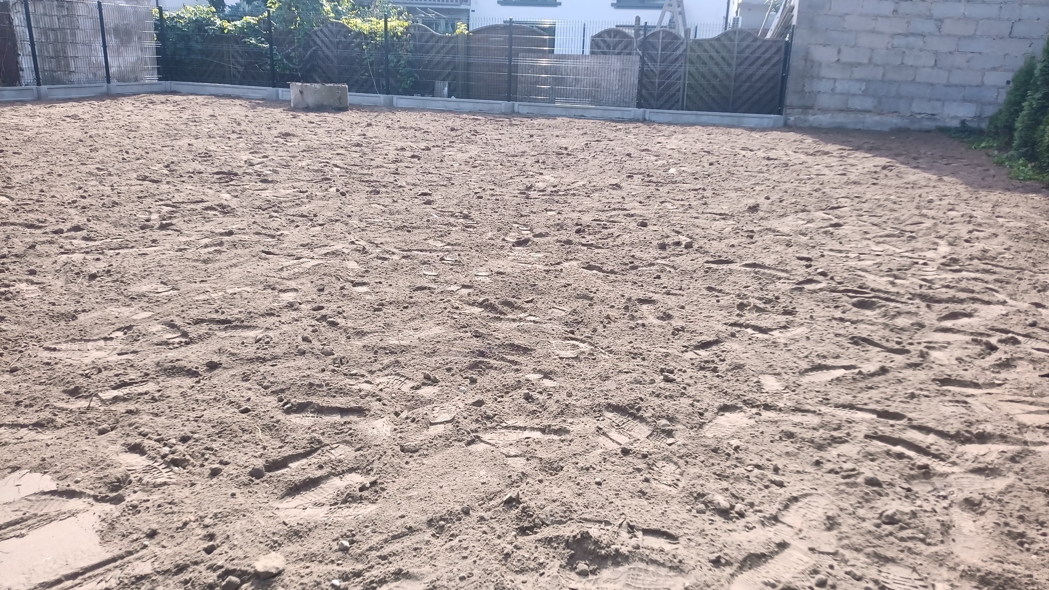 Przygotowany teren pod budowę z widocznymi śladami stóp, ogrodzeniem panelowym i fragmentem budynku w tle, w słoneczny dzień.