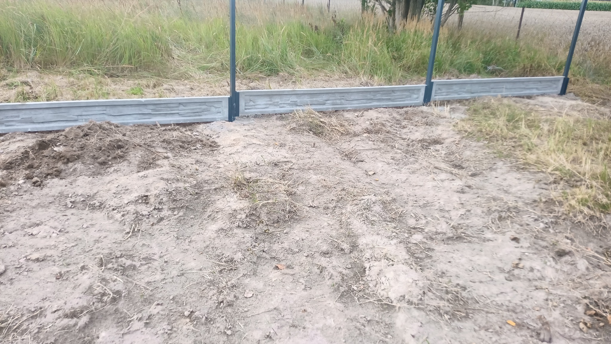 Fragment ogrodzenia z prefabrykatów betonowych imitujących kamień, osadzonych na metalowych słupkach, na tle trawy i ziemi.