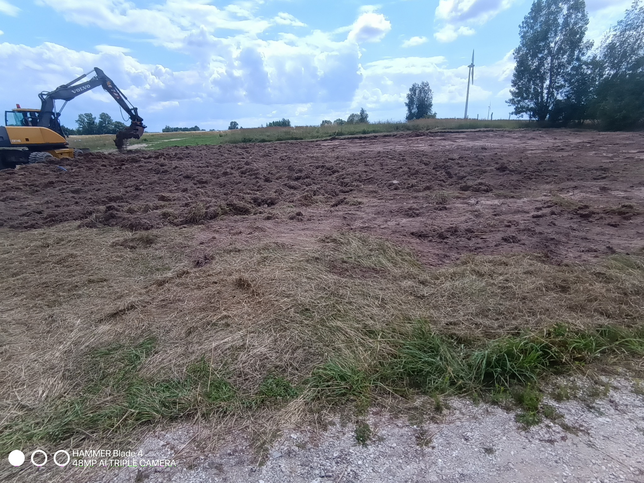 Żółta koparka Volvo na polu z przekopaną ziemią, przygotowując teren pod budowę. W tle widoczne drzewa i turbina wiatrowa pod błękitnym niebem.