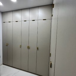 COMFORT ZONE - Beżowa szafa wnękowa z złotymi, okrągłymi uchwytami na tle marmurowej podłogi i białego sufitu z wbudowanym oświetleniem.