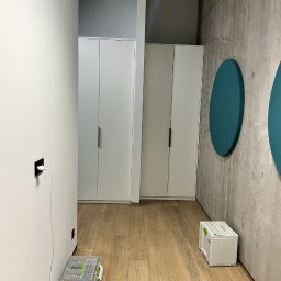 COMFORT ZONE - Montaż szaf wnękowych: widoczne dwie białe szafy z czarnymi uchwytami, skrzynki narzędziowe na podłodze, surowy beton na ścianie z dekoracyjnymi, okrągłymi elementami w kolorze morskim.