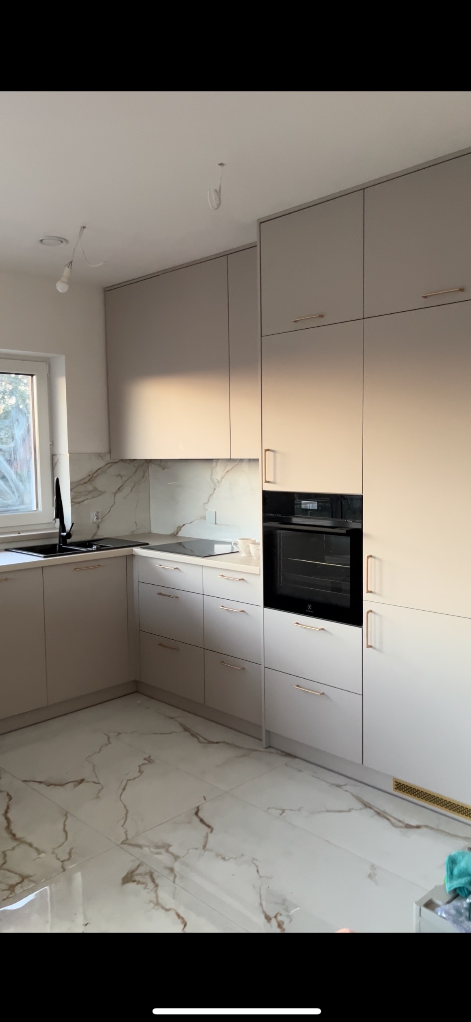 Jasna kuchnia z beżowymi frontami, złotymi uchwytami i marmurowym blatem oraz podłogą, z widocznymi niedokończonymi elementami oświetlenia sufitowego.