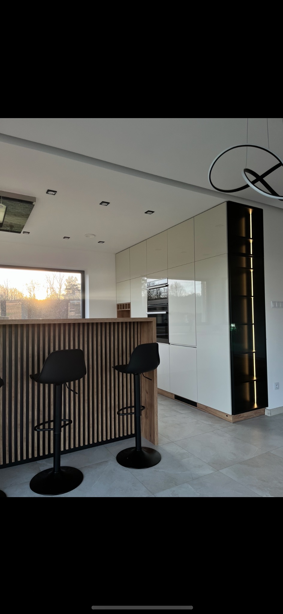Nowoczesna kuchnia z białymi szafkami, czarnym oświetleniem pionowym i drewnianym barem z dwoma czarnymi hokerami na szarych płytkach podłogowych, widok na zachodzące słońce przez okno.