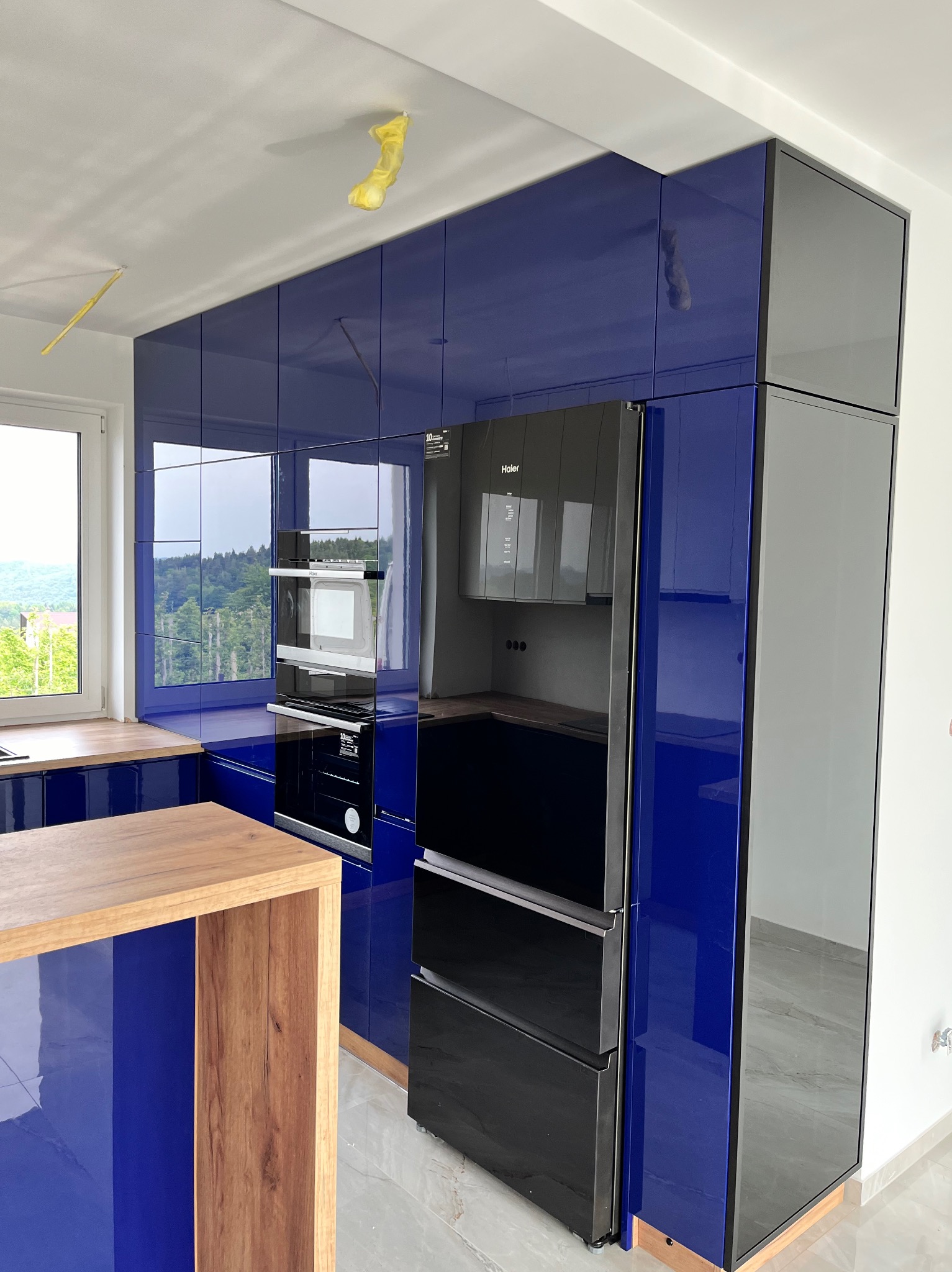 Niebieska, lakierowana zabudowa kuchenna z czarną lodówką Haier i drewnianym blatem, widok na zewnątrz przez okno, folia ochronna na suficie wskazuje na trwający montaż.