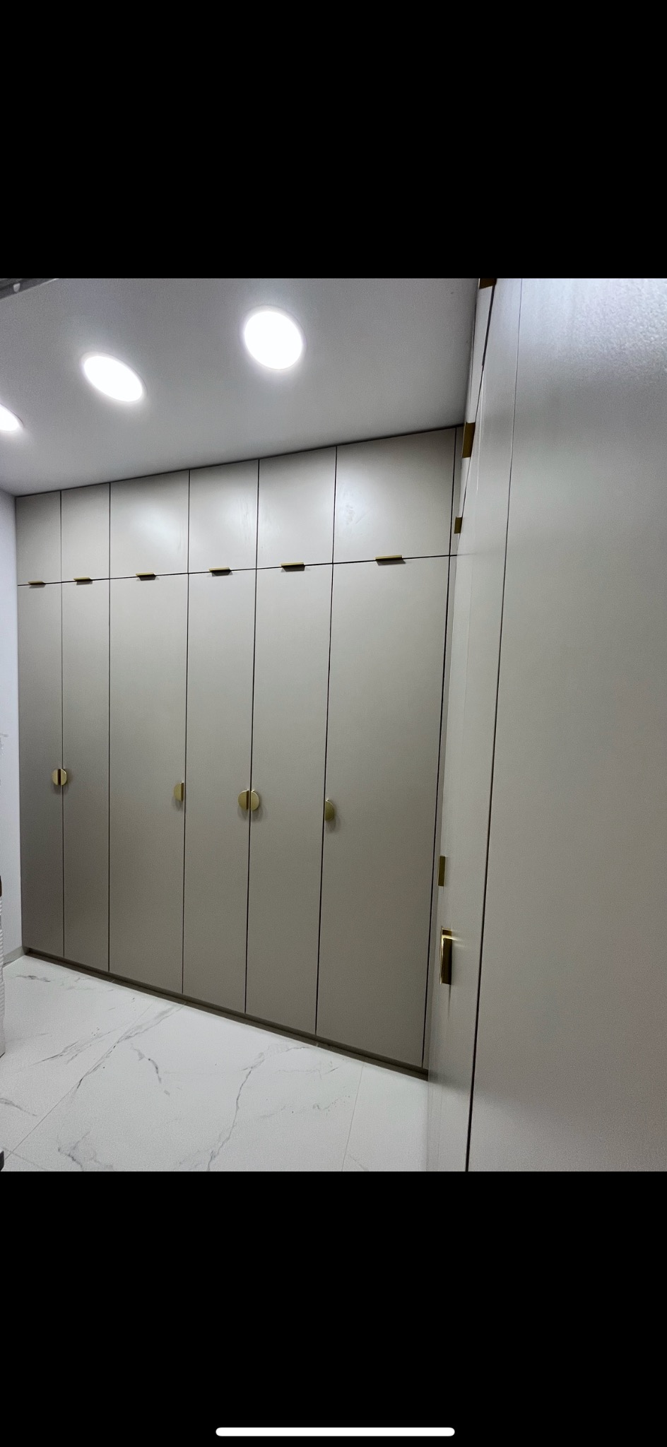 Beżowa szafa wnękowa z złotymi, okrągłymi uchwytami na tle marmurowej podłogi i białego sufitu z wbudowanym oświetleniem.