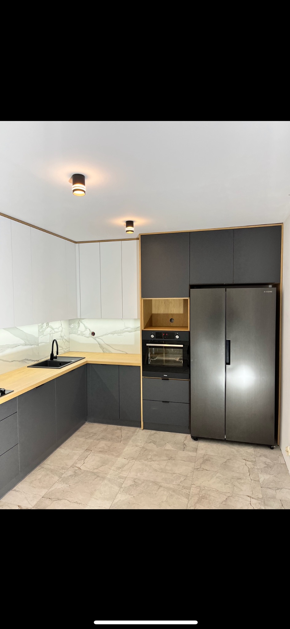 Nowoczesna kuchnia z szarymi i białymi szafkami, drewnianymi blatami, marmurowym backsplashiem i ciemną armaturą, oświetlona dwoma punktowymi lampami sufitowymi.