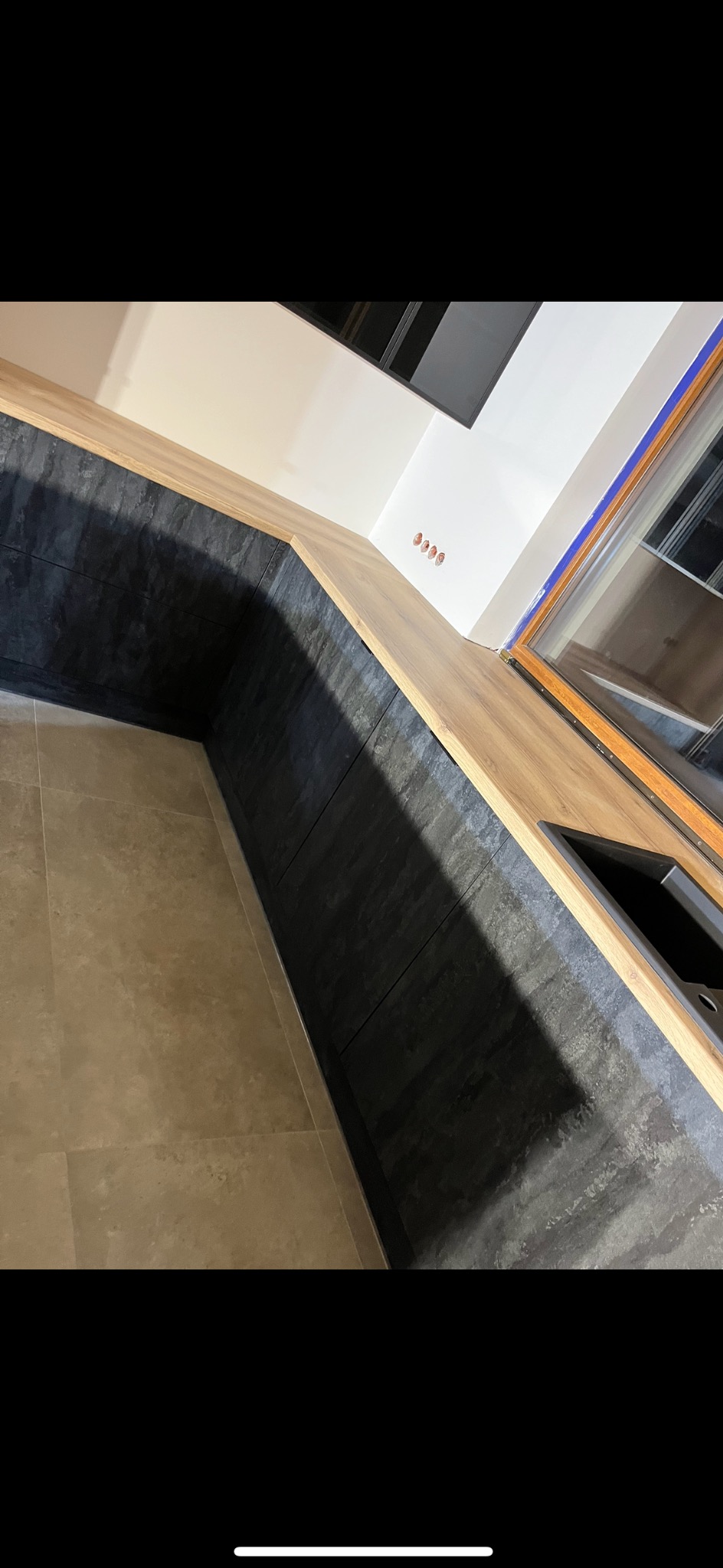 Fragment narożnej zabudowy kuchennej z blatem drewnopodobnym i frontami w kolorze ciemnego grafitu, z widocznym fragmentem zlewu i beżowej podłogi.