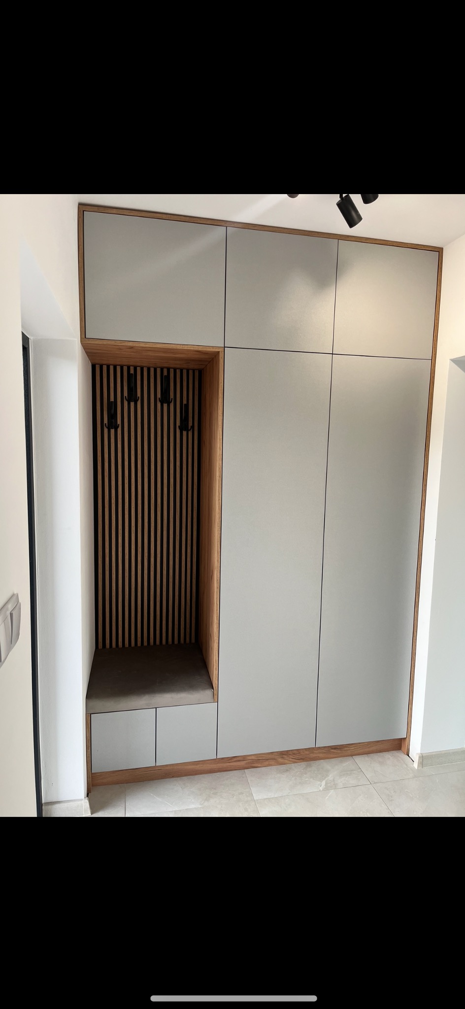 Szara szafa wnękowa z drewnianym wykończeniem, siedziskiem i wieszakami, oświetlona trzema reflektorami sufitowymi.