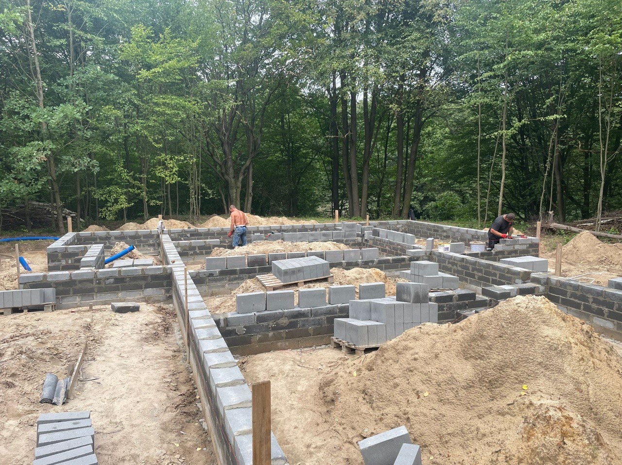 Wykonywanie fundamentów z bloczków betonowych na zalesionej działce, widoczne elementy izolacji poziomej i dwóch pracowników w trakcie murowania.