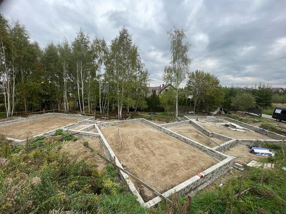 Wykopane i przygotowane podłoże pod kilka fundamentów na działce z widocznymi drzewami i pochmurnym niebem.