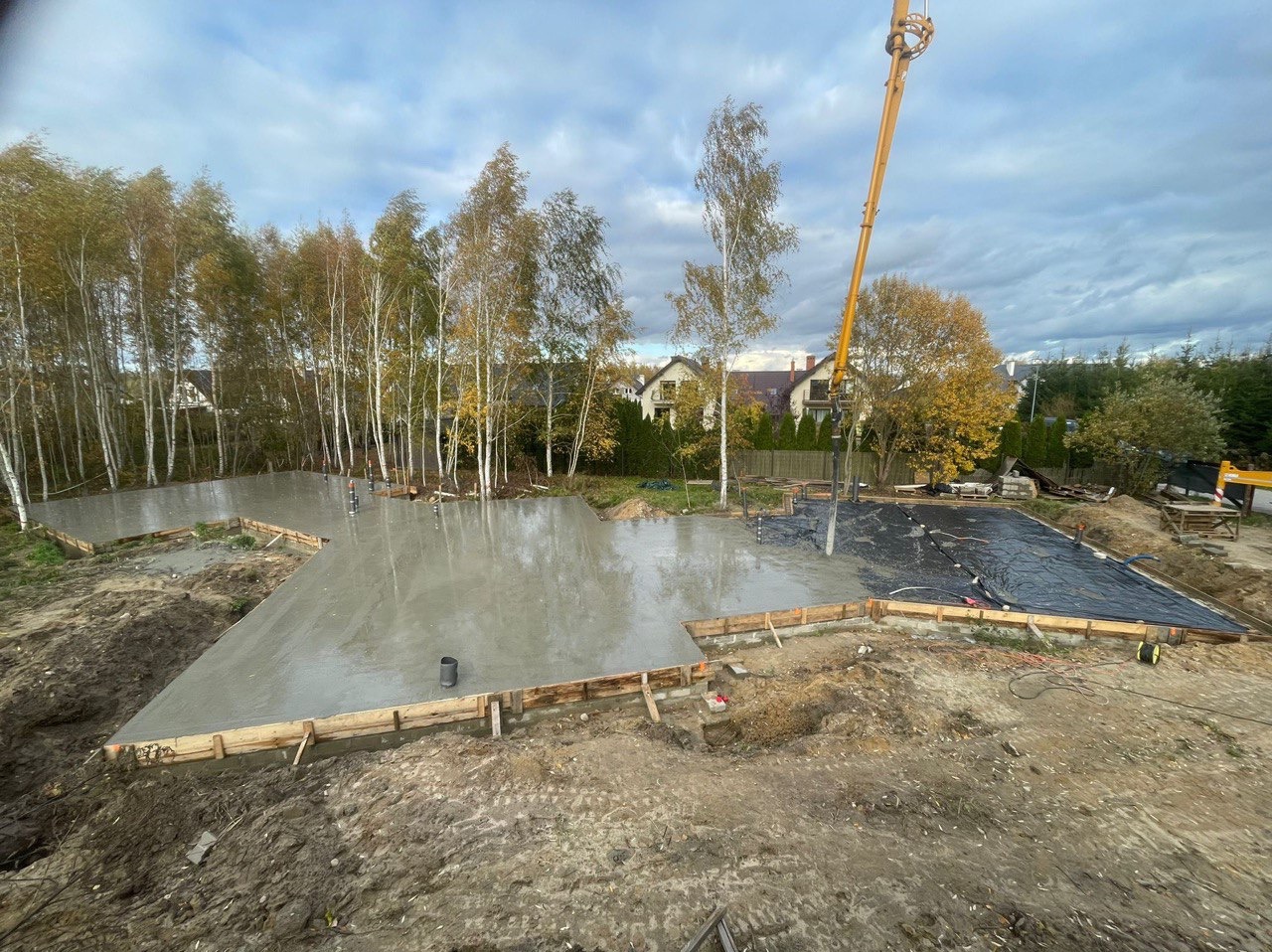 Wylewanie betonu na przygotowany fundament pod budowę domu jednorodzinnego, widoczna pompa do betonu, szalunki drewniane i folia ochronna na części terenu, w tle drzewa i zabudowa mieszkalna.