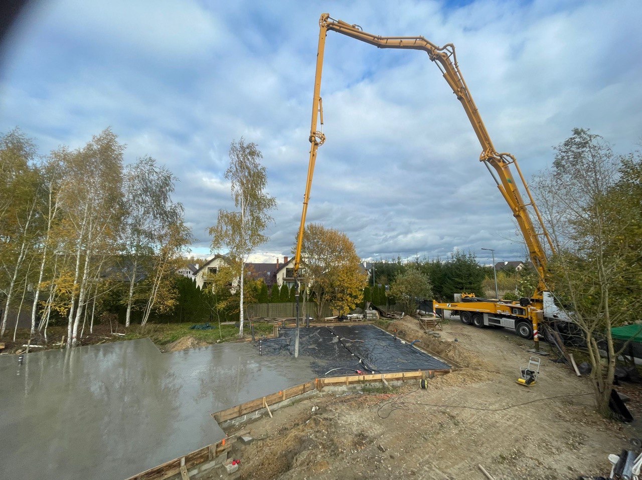 Betonowanie płyty fundamentowej domu jednorodzinnego za pomocą pompy do betonu, widoczne szalunki i folia izolacyjna, w tle drzewa i zabudowa jednorodzinna.