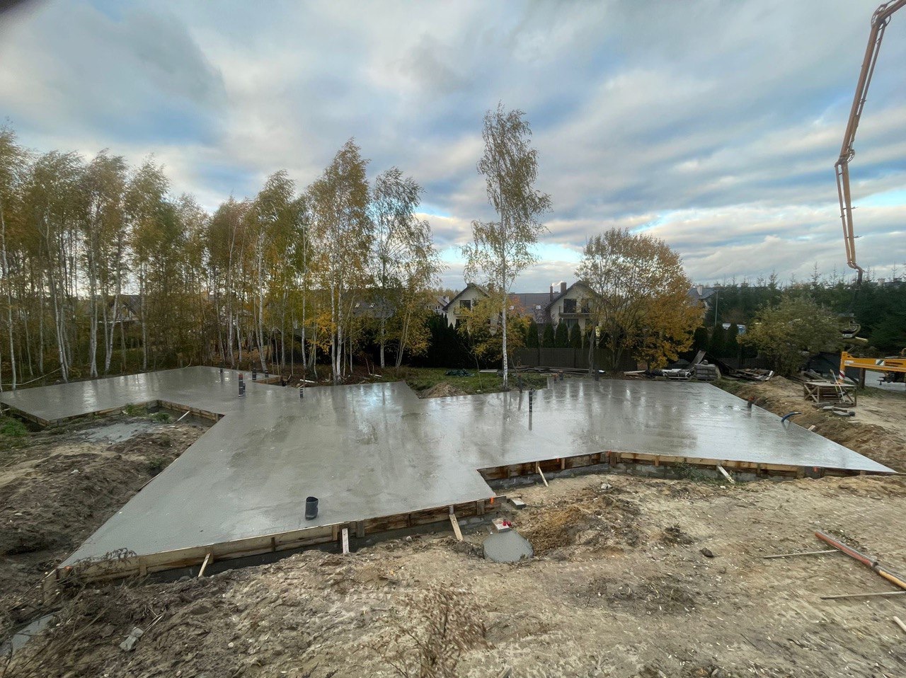 Świeżo wylana, gładka płyta fundamentowa z drewnianym szalunkiem, widoczna pompa do betonu w tle, jesienne drzewa liściaste w otoczeniu.