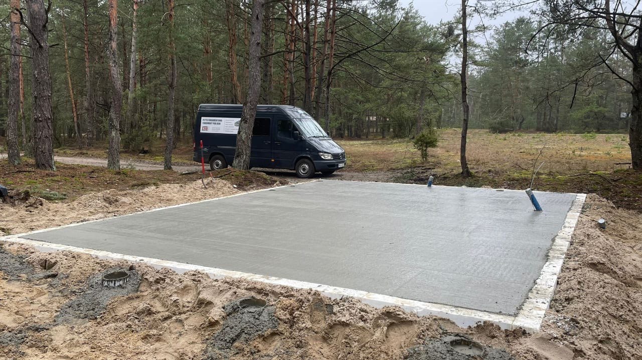 Świeżo wylana betonowa płyta fundamentowa na piaszczystym terenie z widocznymi rurami instalacyjnymi, na tle lasu sosnowego i zaparkowanego busa firmy budowlanej.