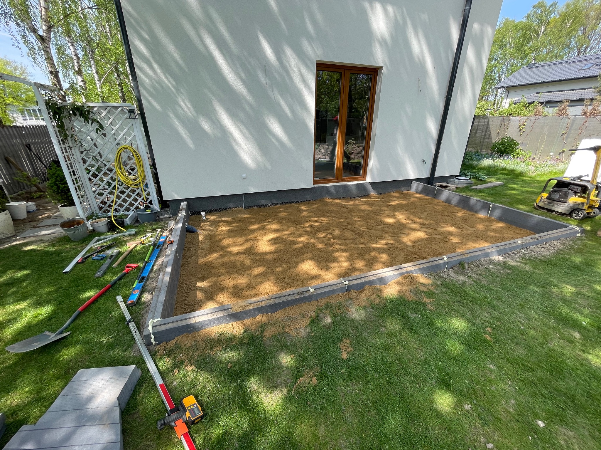 Przygotowany teren pod budowę tarasu z widocznymi narzędziami, poziomnicą laserową i zagęszczarką na trawie.