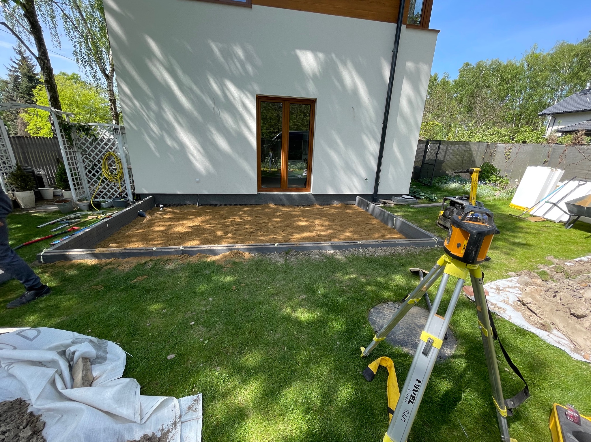 Przygotowanie podłoża pod taras w Warszawie: wyrównany teren z piaskiem w obrębie obrzeży, widoczny niwelator laserowy na statywie, dom z oknem tarasowym w tle.