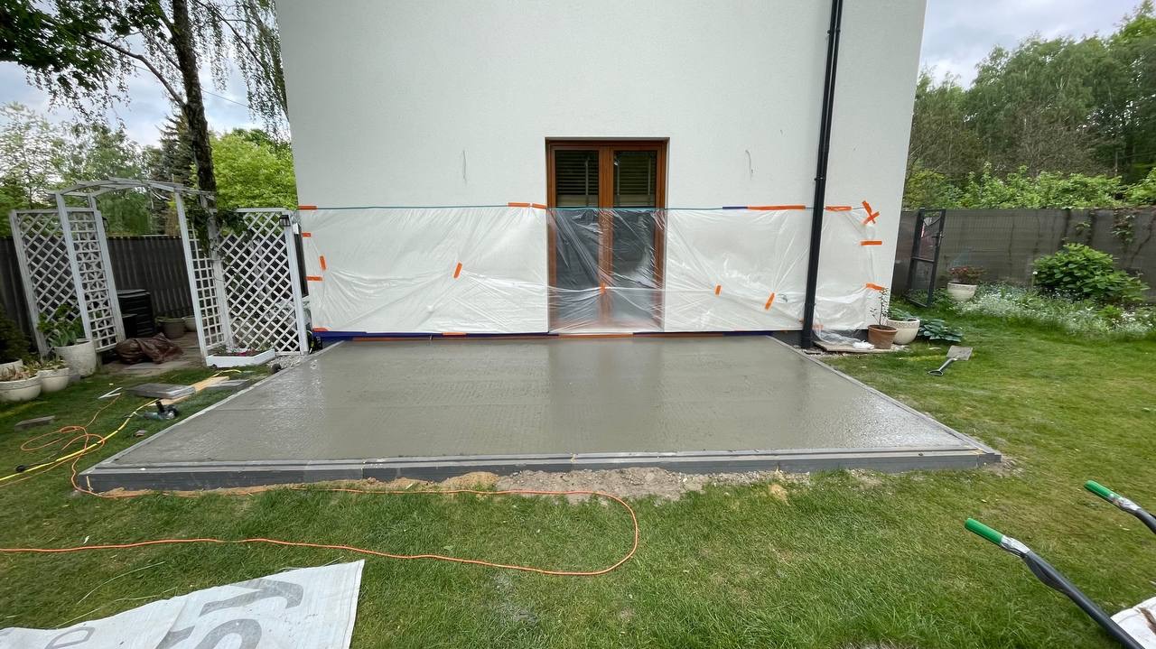 Świeżo wylana, gładka betonowa wylewka tarasu przylegająca do ściany budynku zabezpieczonej folią ochronną, widoczne obrzeża i trawnik, w tle pergola i zieleń.