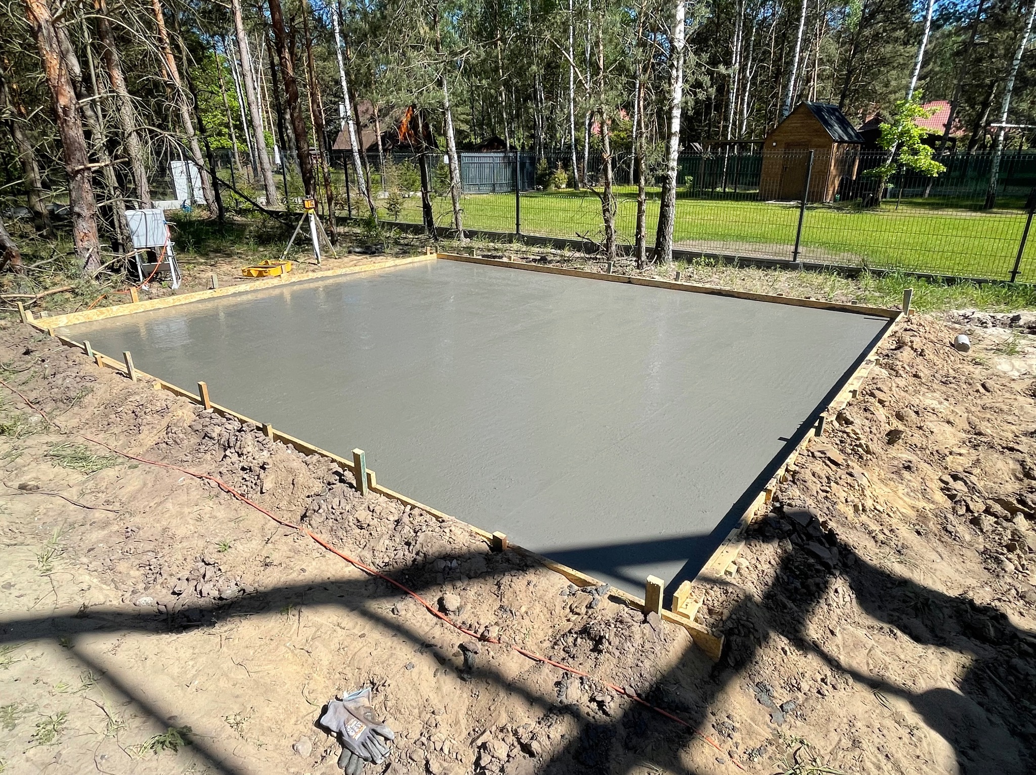 Wylany, świeży beton na przygotowanym placu budowy, otoczony drewnianym szalunkiem, z niwelatorem laserowym w tle i lasem w oddali, gotowy do dalszych prac.