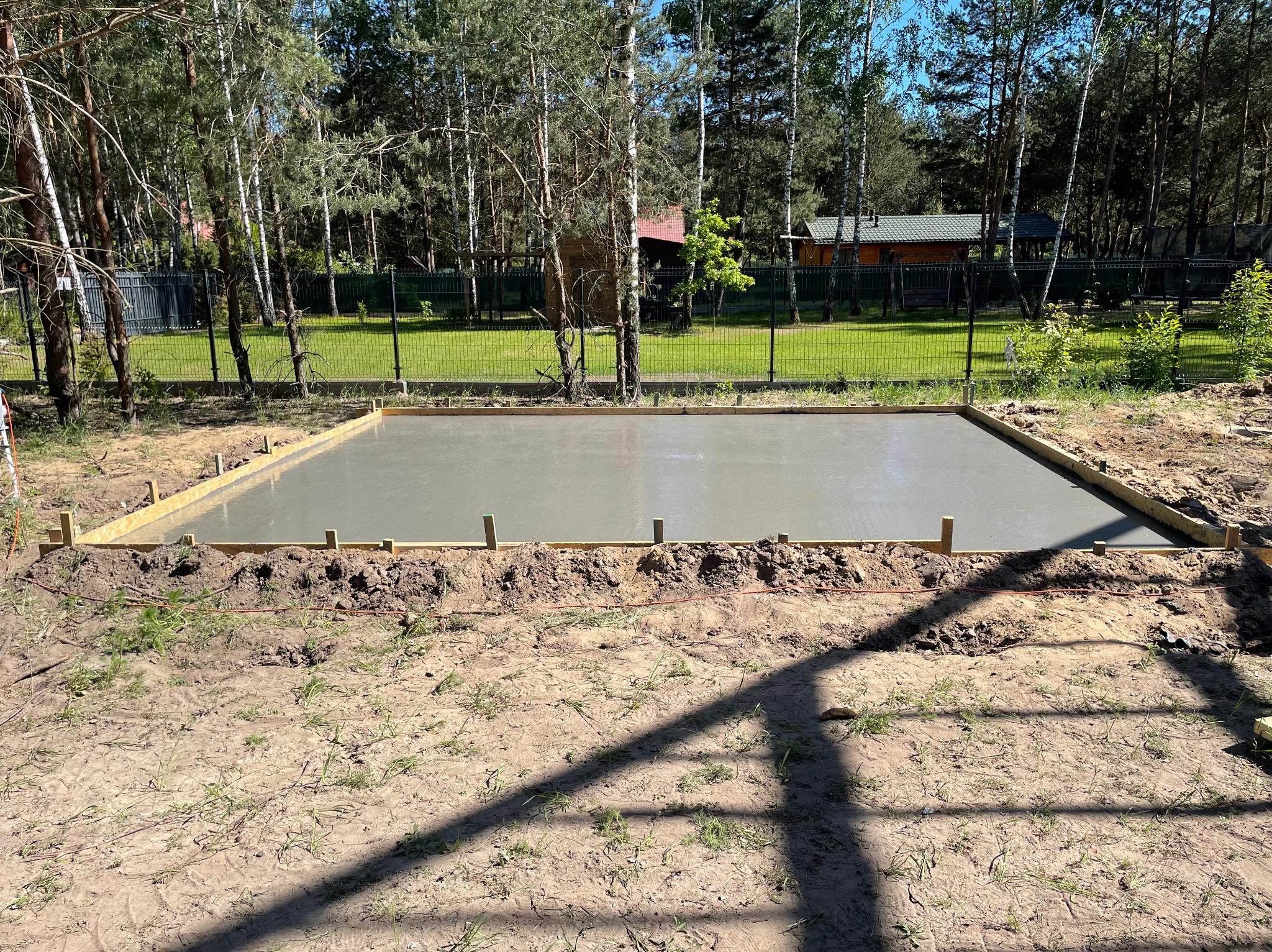 Świeżo wylany betonowy fundament pod budowę domu, otoczony drewnianym szalunkiem i widokiem na zalesiony teren w tle.
