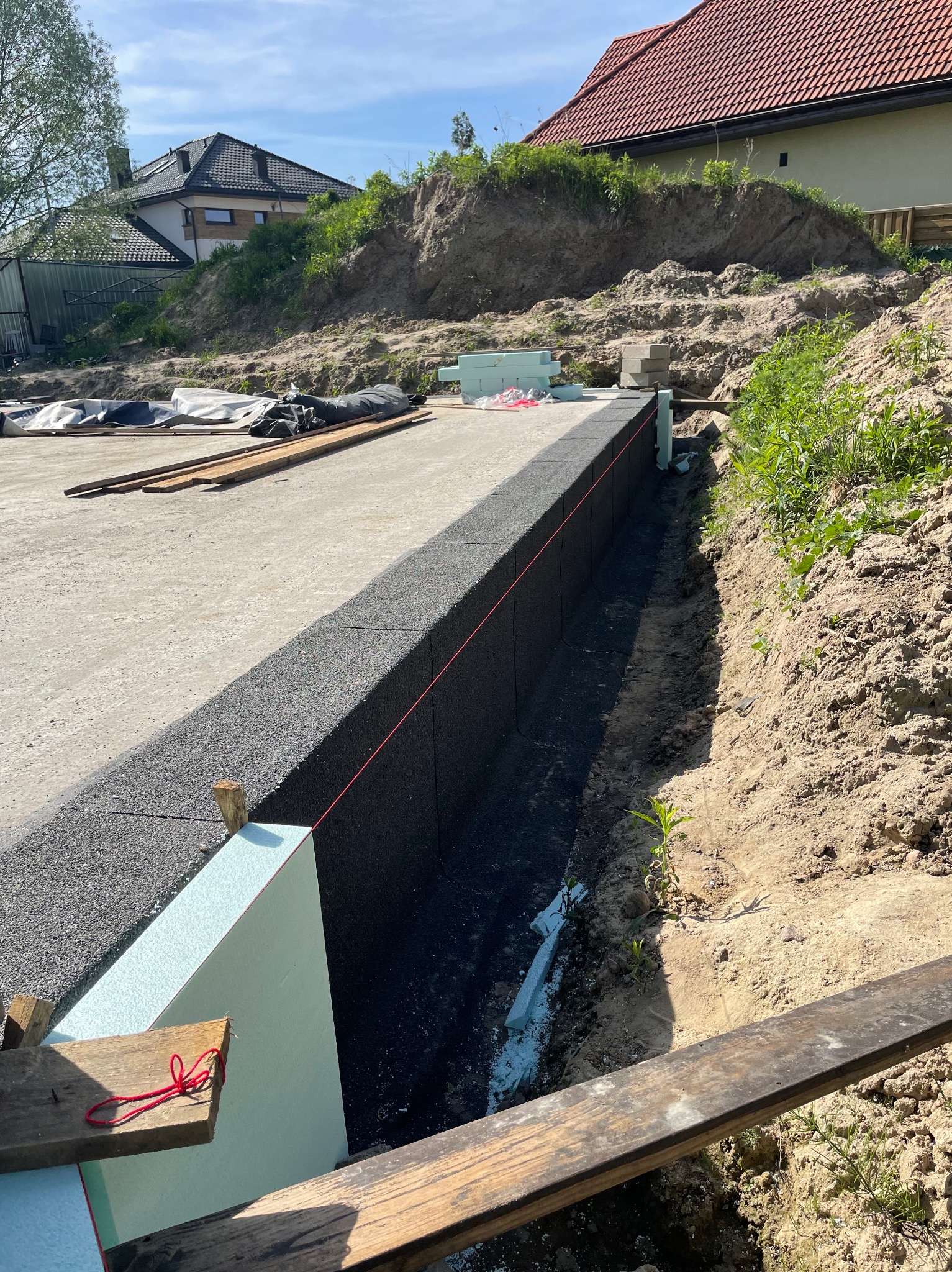 Przygotowanie do wylewki fundamentów: widoczne szalunki z izolacją termiczną, czarna izolacja pionowa, wytyczona linia pozioma, teren wyrównany z widoczną skarpą ziemi w tle.