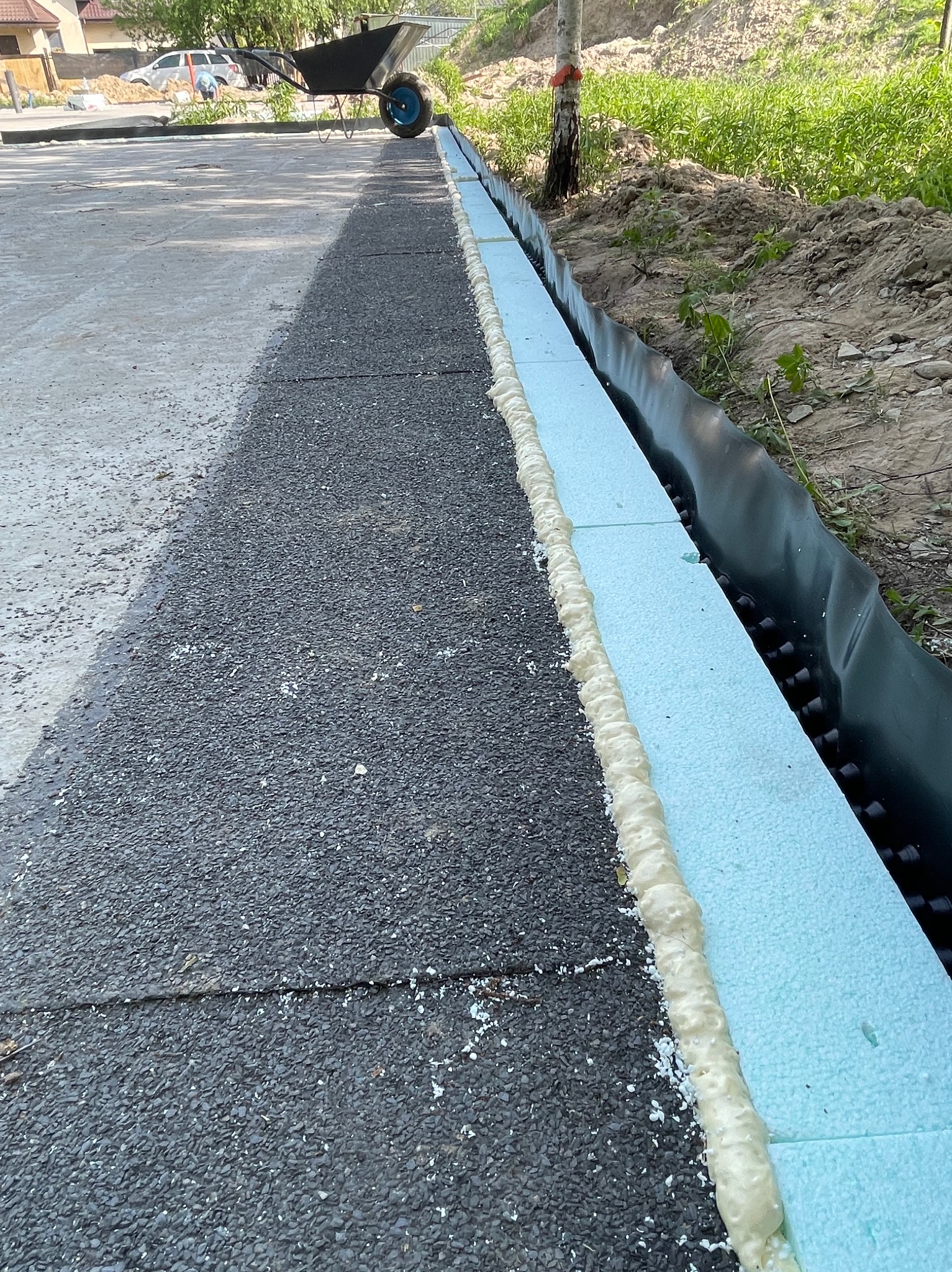 Przygotowanie podłoża pod drogę: warstwa izolacyjna z niebieskiego styropianu zabezpieczona pianką montażową, czarna folia drenażowa, widoczny grunt, w tle taczka.