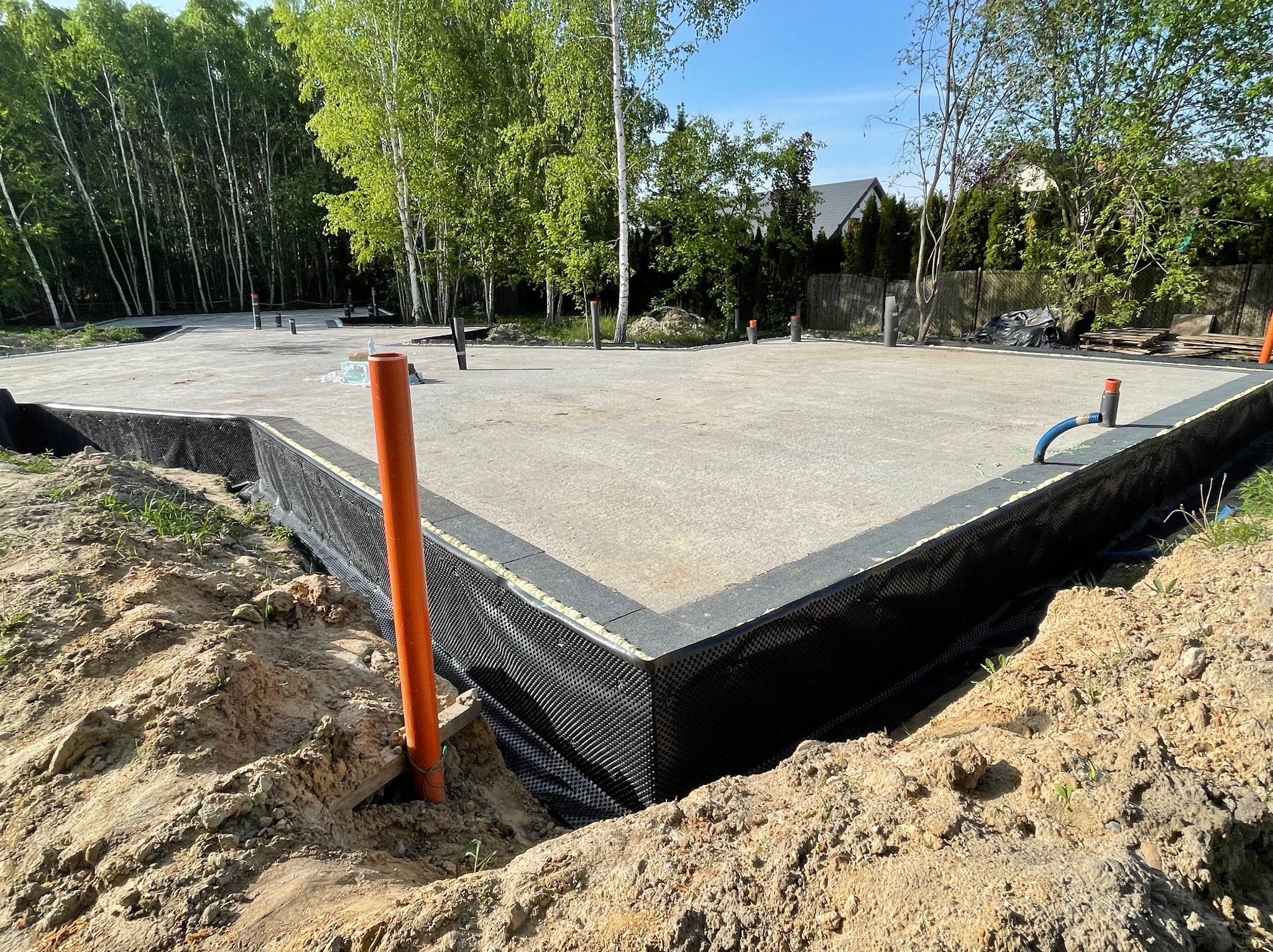 Wylany betonowy fundament płyty, zabezpieczony czarną folią izolacyjną i styropianem, z wystającymi pionowymi rurami PCV w kolorze pomarańczowym, otoczony ziemią i zielenią, w tle drzewa i dom.