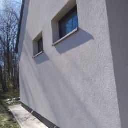 Ściana budynku z oknami po nałożeniu jasnoszarego tynku strukturalnego. Widoczne refleksy światła i cień. Na dole chodnik z kostki brukowej i przewody.