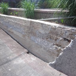 RENO-GŁADŹ MARCIN BRZOZOWSKI - Nierówna, betonowa ściana oporowa z ubytkami i śladami napraw, przylegająca do chodnika z kostki. Na górze roślinność wzdłuż ściany.