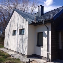 RENO-GŁADŹ MARCIN BRZOZOWSKI - Nowoczesny dom jednorodzinny z białą elewacją i grafitowym dachem z blachy na tle drzew i błękitnego nieba. Widoczne okna z grafitową stolarką i rynny.