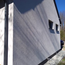 RENO-GŁADŹ MARCIN BRZOZOWSKI - Ściana budynku z szarą elewacją o strukturze baranka, widoczne dwa okna i rynna. Na dole czarny pas. Na chodniku leży biały kabel.