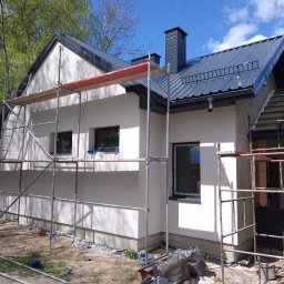 RENO-GŁADŹ MARCIN BRZOZOWSKI - Dom jednorodzinny z rusztowaniem podczas prac elewacyjnych. Widoczna świeżo nałożona, jasna warstwa tynku. Ujęcie z boku, w tle drzewa i błękitne niebo.