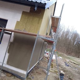 RENO-GŁADŹ MARCIN BRZOZOWSKI - Ocieplanie domu: fragment elewacji z widocznym styropianem, wełną mineralną i nałożonym tynkiem. Rusztowanie przy ścianie. Prace w toku, widoczne materiały budowlane.