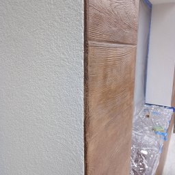 RENO-GŁADŹ MARCIN BRZOZOWSKI - Dekoracyjny narożnik ściany z efektem drewna, sąsiadujący z białą, chropowatą powierzchnią. Widoczne zabezpieczenia folią malarską. Detal wykończenia wnętrza.