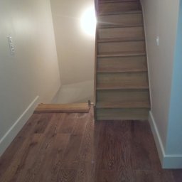 RENO-GŁADŹ MARCIN BRZOZOWSKI - Drewniane schody i podłoga w Hucisku. Jasne ściany, minimalistyczny design. Widoczne detale wykończenia i naturalna tekstura drewna. Ujęcie z dołu, podkreślające wysokość.