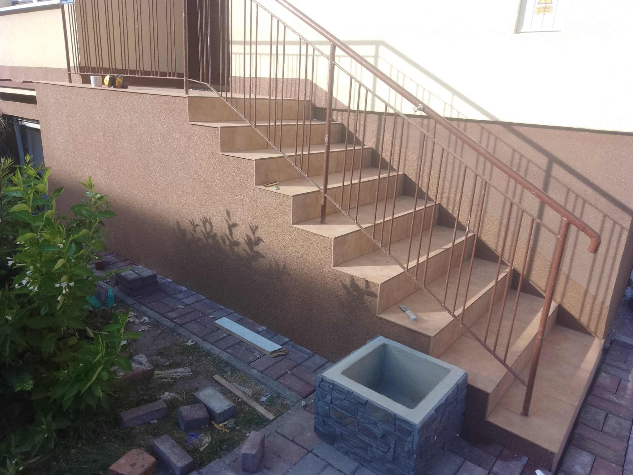 Zewnętrzne schody z brązową balustradą i wykończone płytkami, prowadzące do budynku z elewacją w odcieniach brązu. Widoczne elementy ogrodu i kostka brukowa.