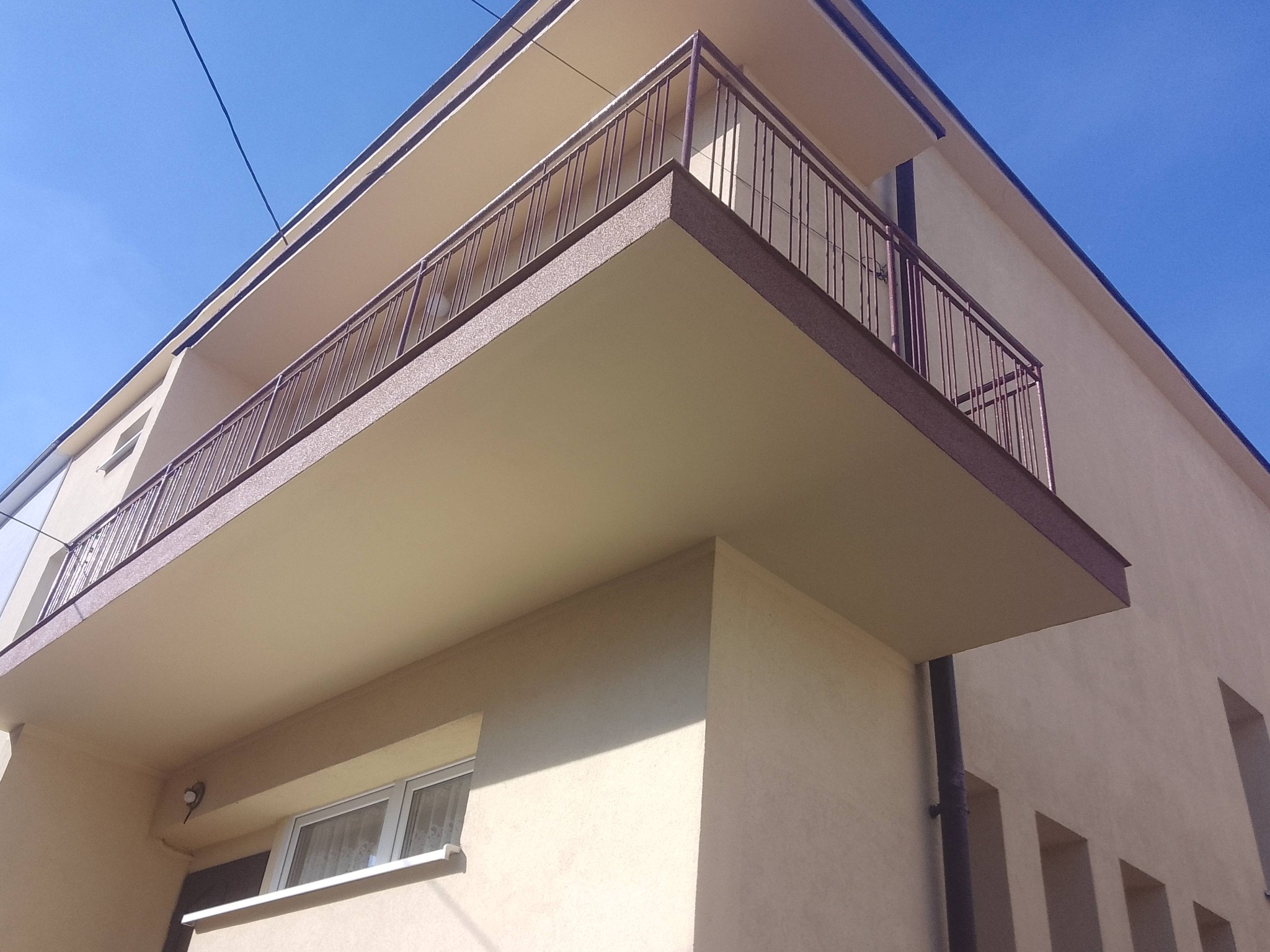 Jasny budynek z balkonem o brązowych barierkach, widziany z dołu pod kątem. Świeżo pomalowana elewacja i błękitne niebo w tle.