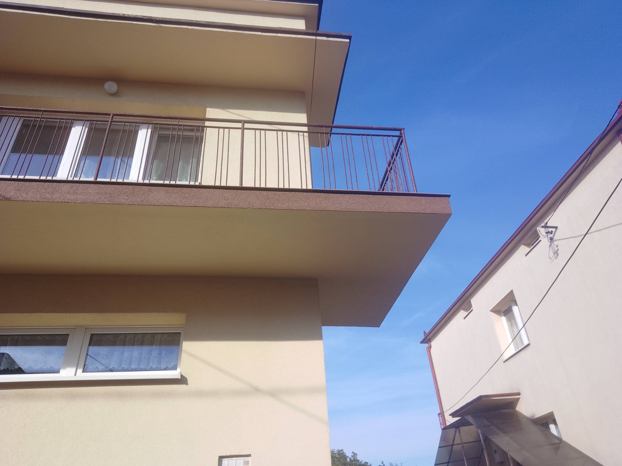 Jasna elewacja budynku z balkonem i oknem, widok z dołu na błękitne niebo. Minimalistyczna architektura, spokojna kolorystyka, wyraźne linie i kąty.