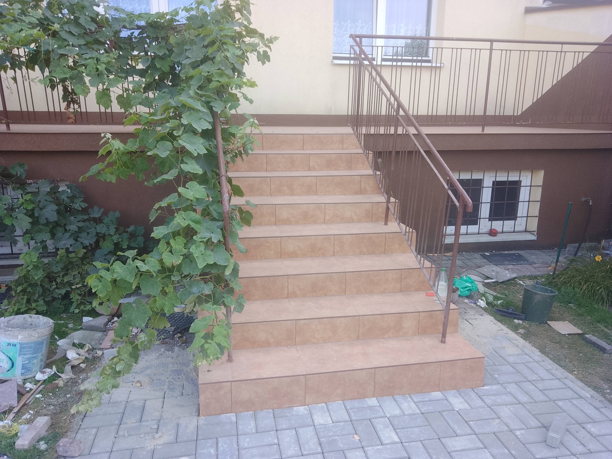 Wyłożone kafelkami schody zewnętrzne z metalową barierką, obok roślina pnąca. Widoczne okna budynku w tle. Jasne płytki schodowe i szara kostka brukowa.