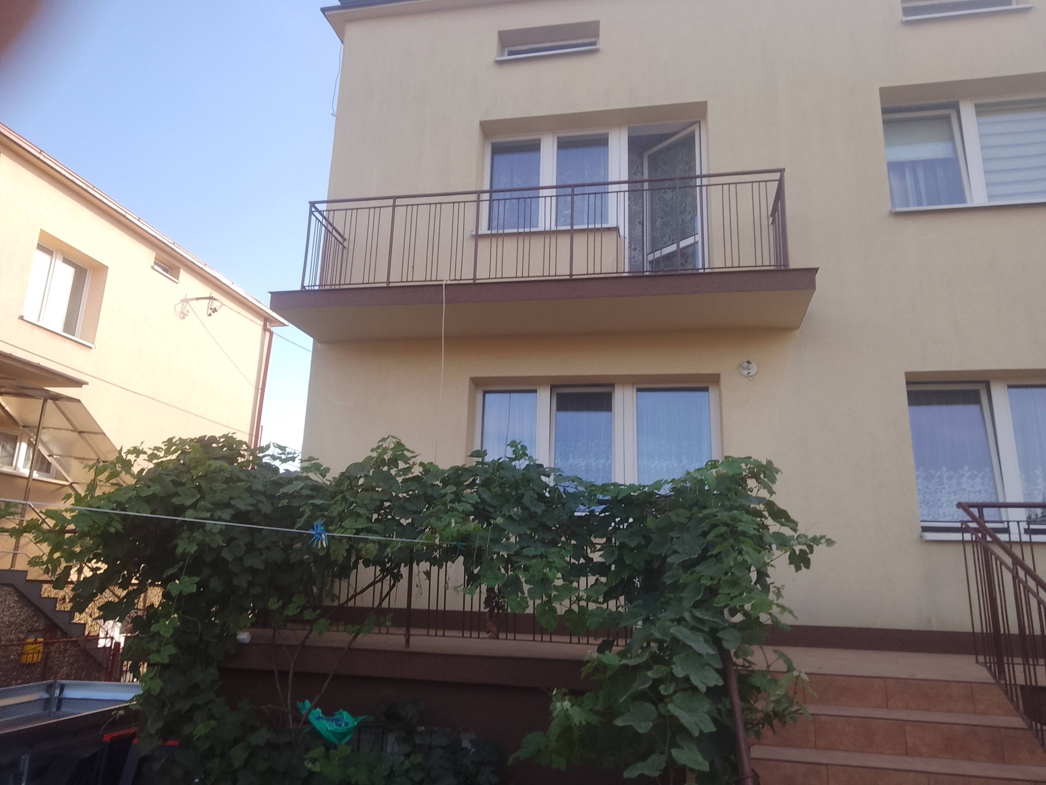 Dwupiętrowy dom z beżową elewacją i balkonem z metalową balustradą. Roślinność pnąca się po balkonie na parterze. Częściowy widok sąsiedniego budynku.