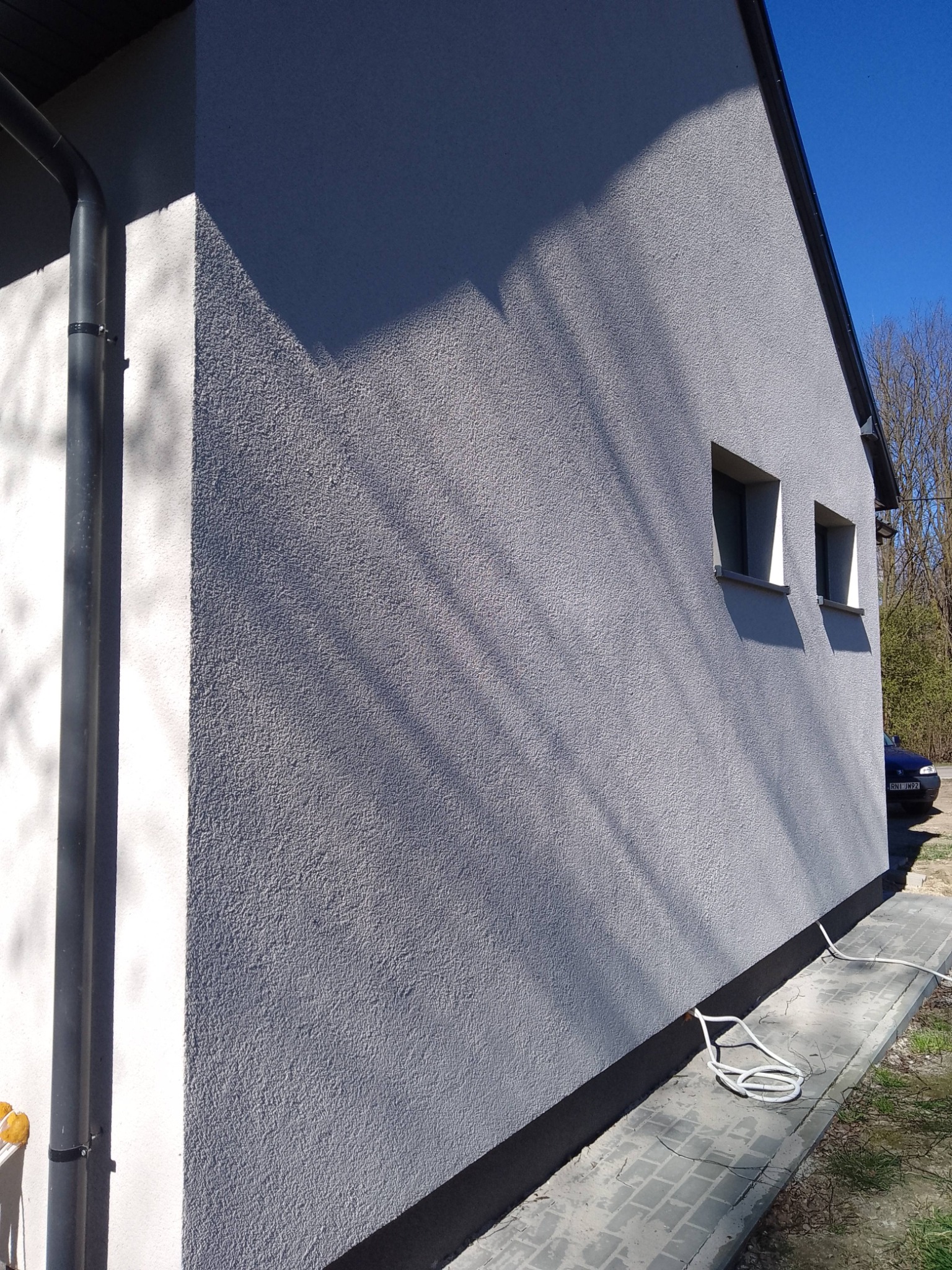 Ściana budynku z szarą elewacją o strukturze baranka, widoczne dwa okna i rynna. Na dole czarny pas. Na chodniku leży biały kabel.