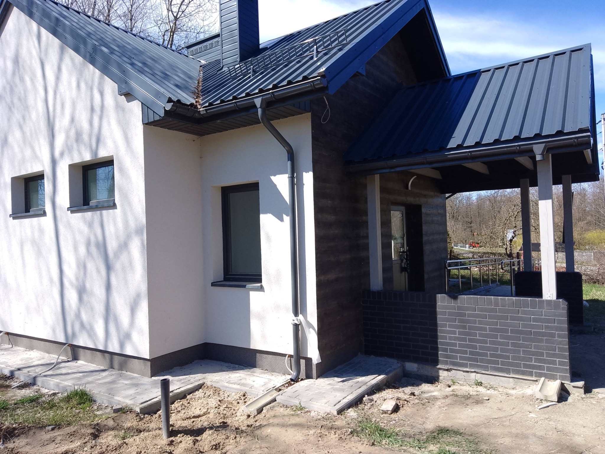 Nowoczesny dom jednorodzinny z białą elewacją i grafitowym dachem z blachy. Widoczne okna, rynny i fragment tarasu z ceglaną podmurówką. Ujęcie w słoneczny dzień.