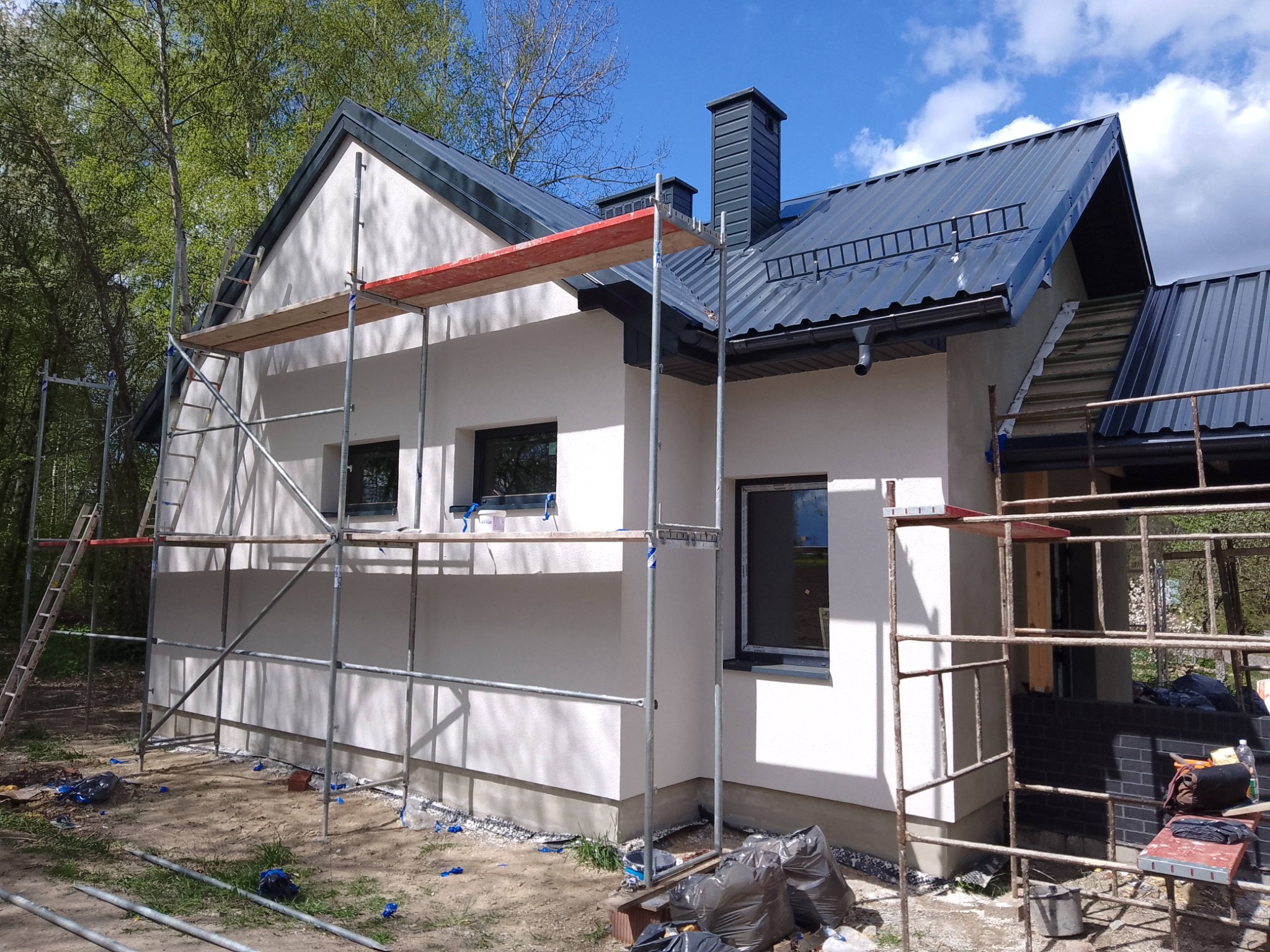 Dom jednorodzinny z rusztowaniem podczas prac elewacyjnych. Widoczna świeżo nałożona, jasna warstwa tynku. Ujęcie z boku, w tle drzewa i błękitne niebo.