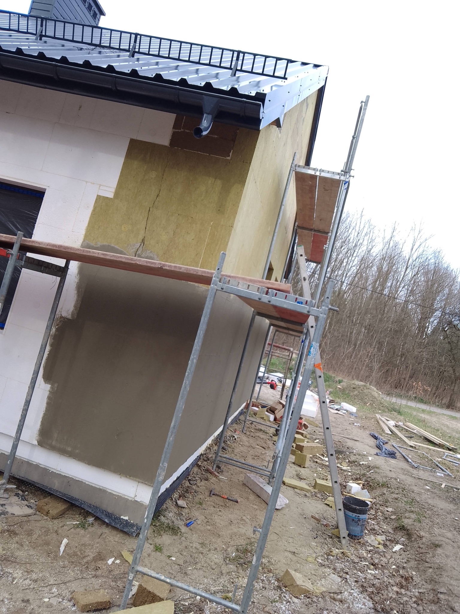Ocieplanie domu: fragment elewacji z widocznym styropianem, wełną mineralną i nałożonym tynkiem. Rusztowanie przy ścianie. Prace w toku, widoczne materiały budowlane.