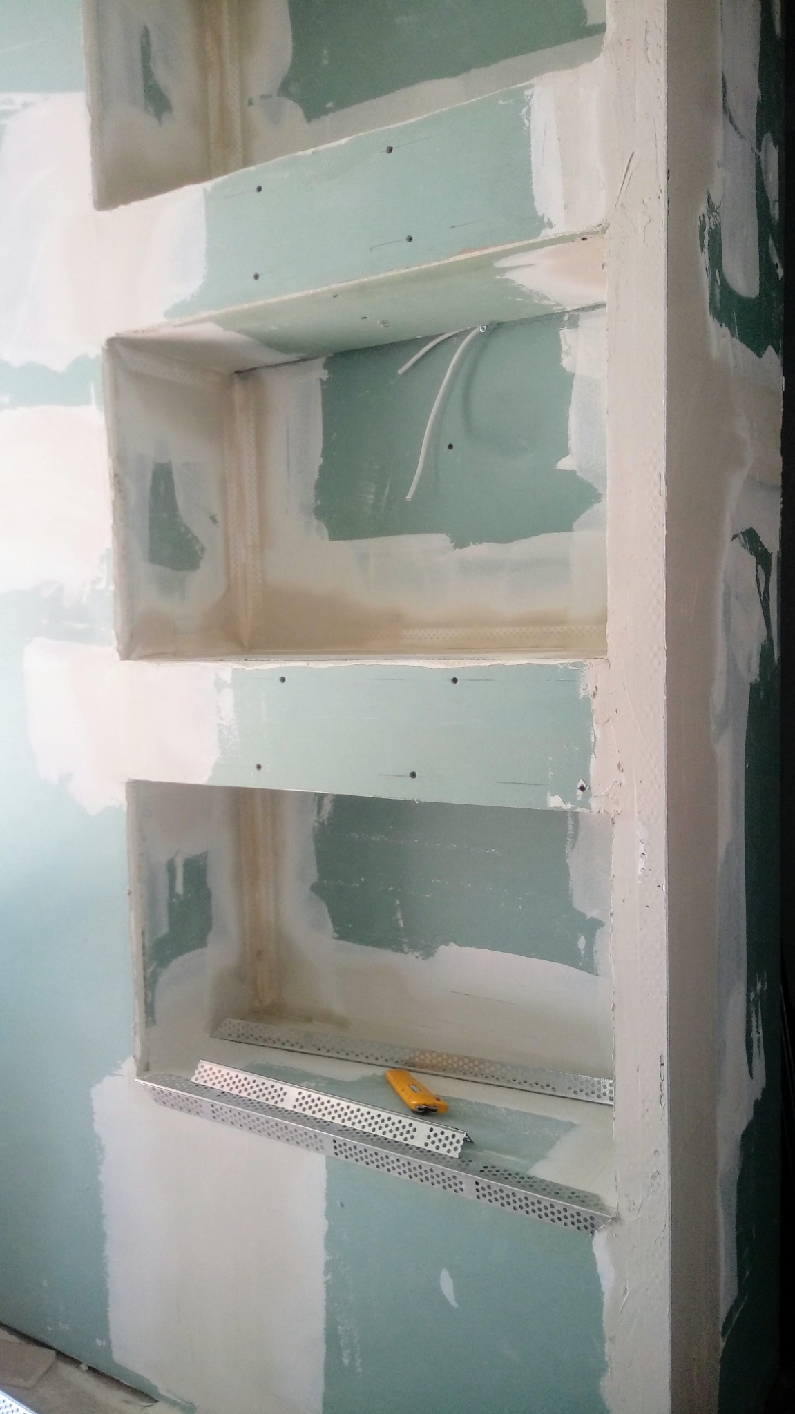 Narożnikowo-półkowy detal wnęki ściennej z płyt gipsowo-kartonowych, częściowo wyszpachlowany, z widocznym nożykiem do tapet na dolnej półce.