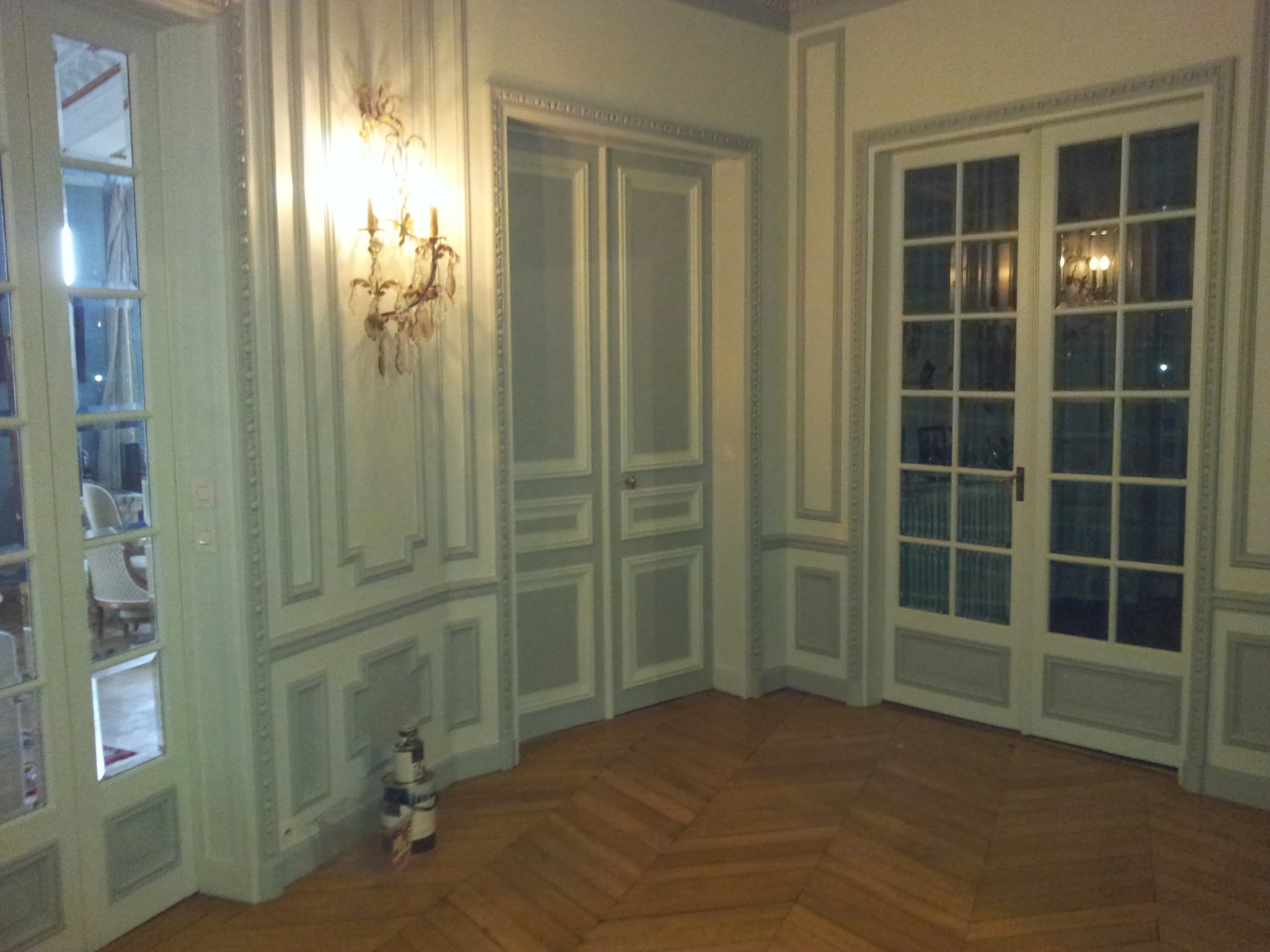 Eleganckie wnętrze z parkietem w jodełkę, ozdobnymi panelami ściennymi i kryształowym kinkietem. Widoczne drzwi balkonowe i drzwi wewnętrzne z dekoracyjnymi listwami.