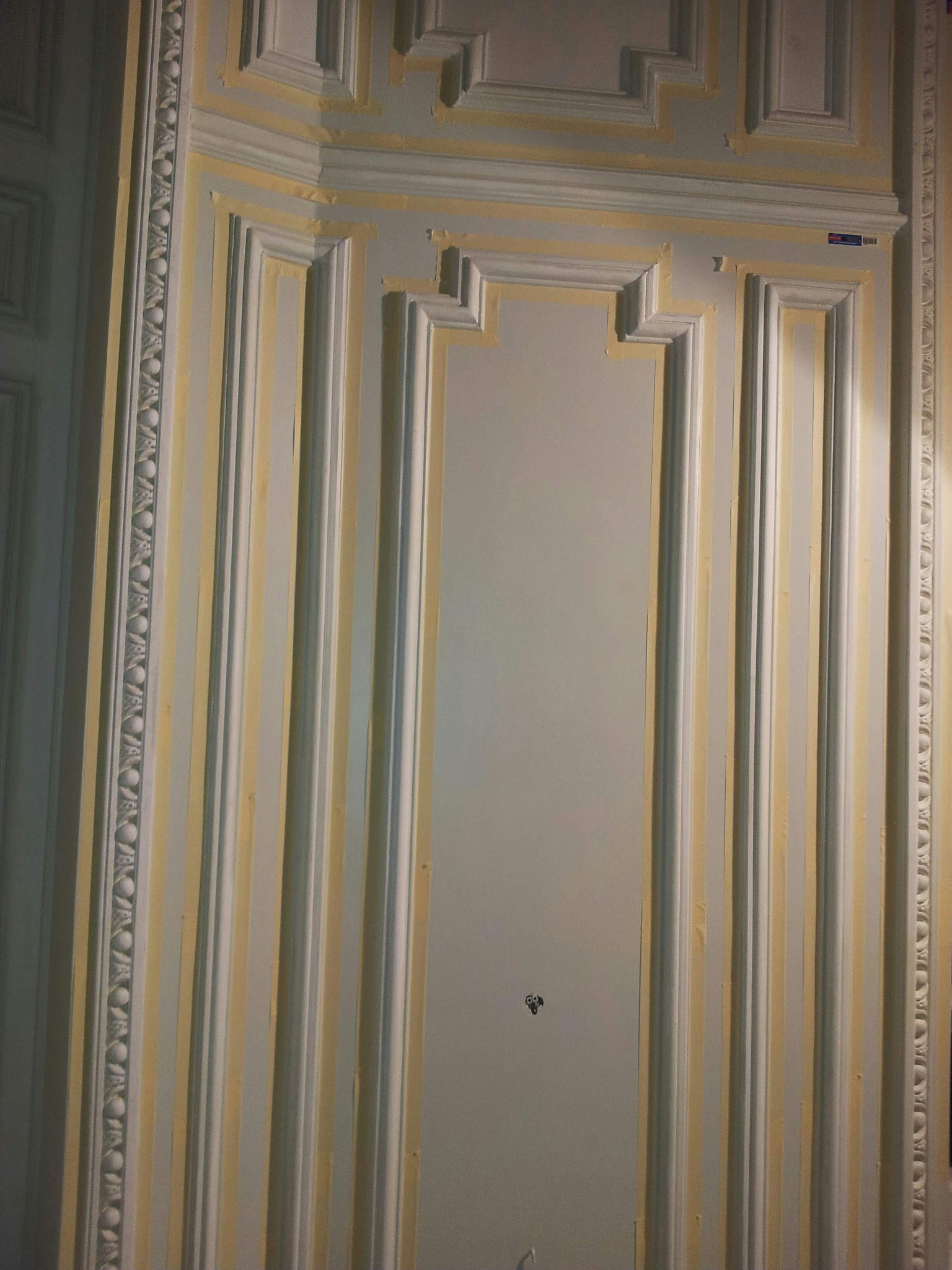 Dekoracyjna ściana z listwami i taśmą malarską, przygotowana do malowania. Widoczne zdobienia i detale architektoniczne. Oświetlenie podkreśla fakturę.