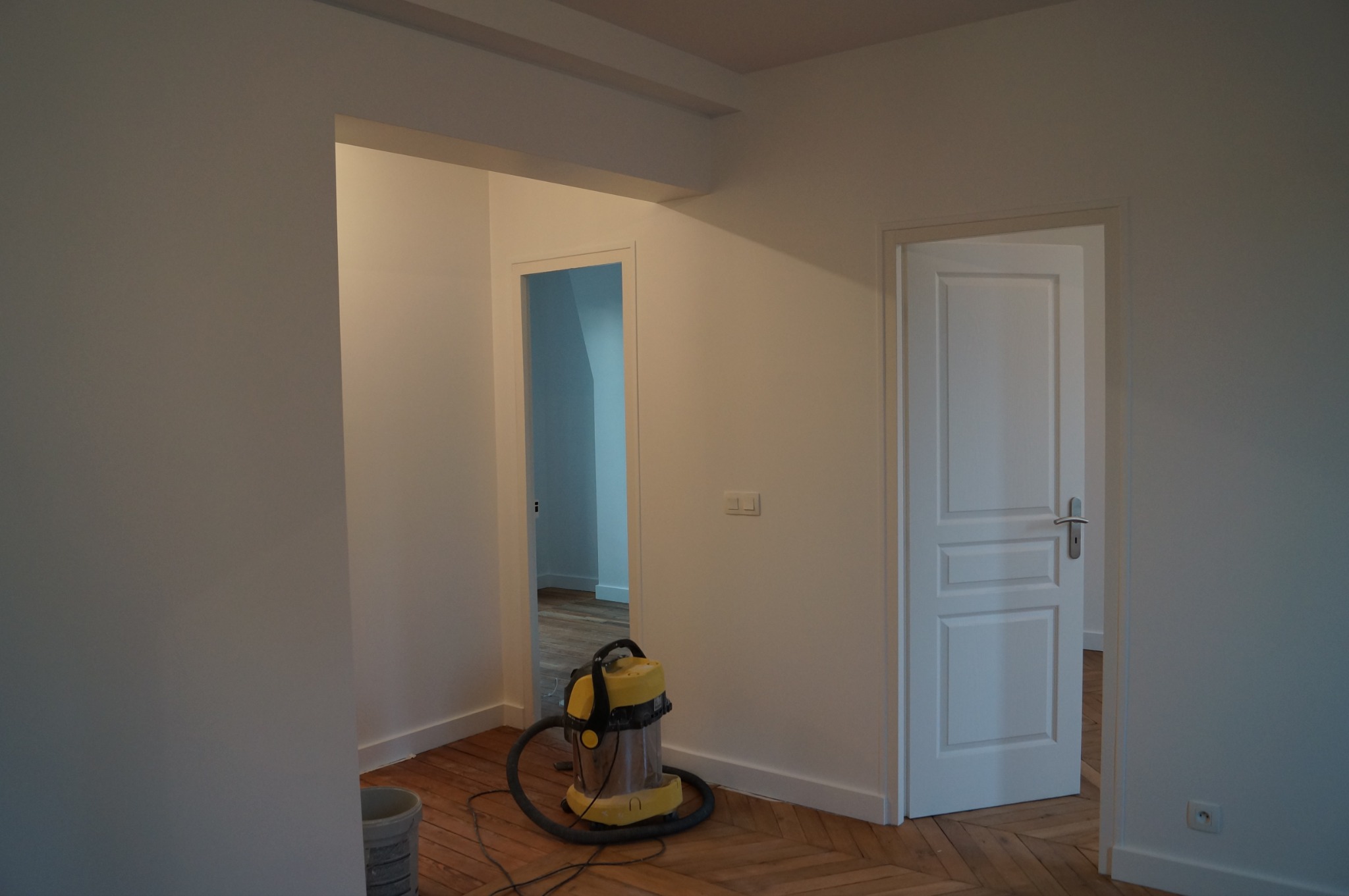 Wnętrze po remoncie: świeżo pomalowane ściany w odcieniach bieli, drewniana podłoga w jodełkę, białe drzwi i odkurzacz na podłodze. Widok na sąsiednie pomieszczenie.