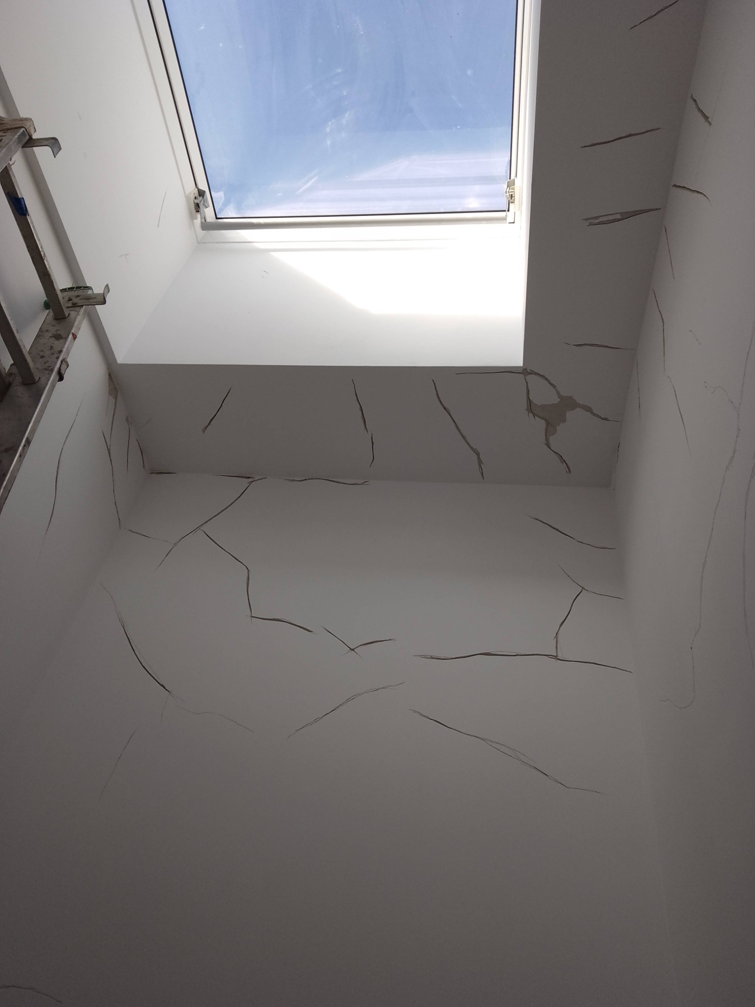 Białe ściany z pęknięciami pod oknem dachowym, widoczny fragment drabiny. Uszkodzona powierzchnia przed remontem, wymagająca szpachlowania i malowania.