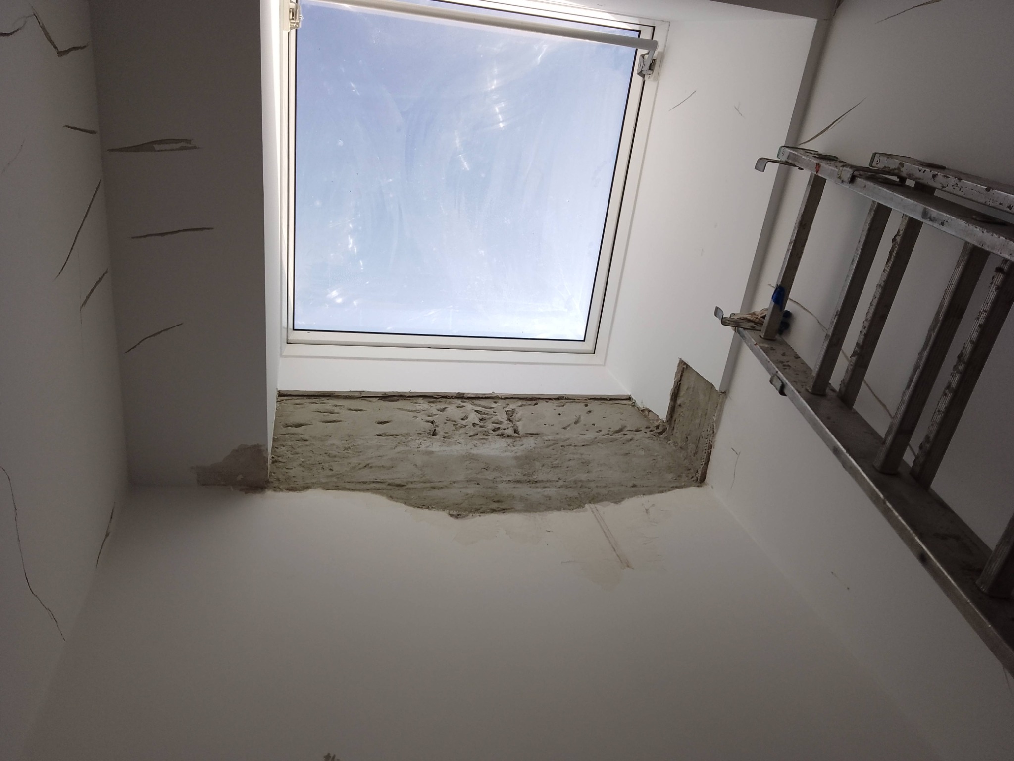 Wnętrze w trakcie remontu: ściany z pęknięciami, fragment ściany bez tynku przy oknie dachowym, drabina oparta o ścianę. Widok na błękitne niebo przez okno.