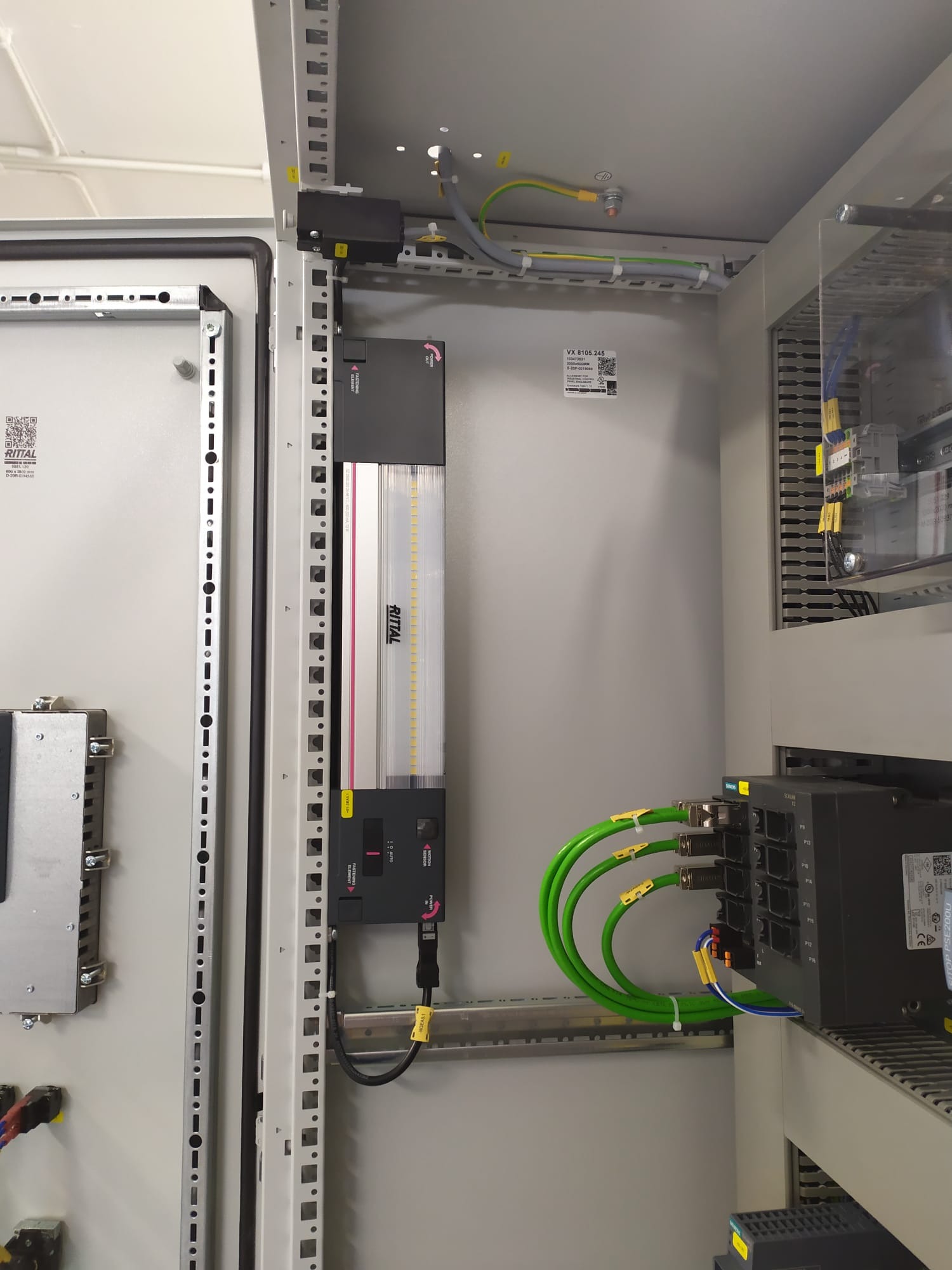 Wnętrze szafy sterowniczej Rittal z zainstalowanym oświetleniem LED, modułami sterującymi i okablowaniem z zielonymi przewodami Ethernet, widoczne oznaczenia i etykiety na elementach.